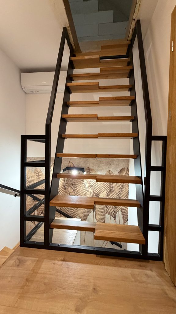 Realizacja schodów na konstrukcji stalowej z drewnianymi stopniami i czarną balustradą. Projekt dopasowany do wnętrza mieszkalnego z naciskiem na estetykę, trwałość i lekkość wizualną.